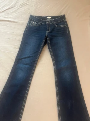 Mörkblå bootcut jeans - Säljer ett par mörkblå bootcut jeans med vita kontrastsömmar och låg midja. Jeansen har klassiska fem fickor och en liten ficka med knappdetalj fram. 