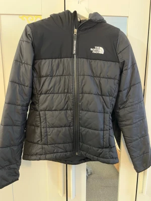 Svart pufferjacka från The North Face - Snygg svart pufferjacka från The North Face med huva och dragkedja framtill. Jackan har quiltad design, två sidofickor och logga på bröstet. Perfekt för kalla dagar och håller dig varm med sin vadderade insida.