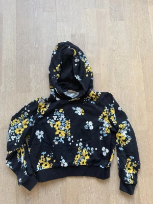 Svart hoodie med blommigt mönster - Snygg svart hoodie med huva och blommigt mönster i gult, vitt och blått. Tröjan har ribbade muddar vid ärmslut och nederkant. 