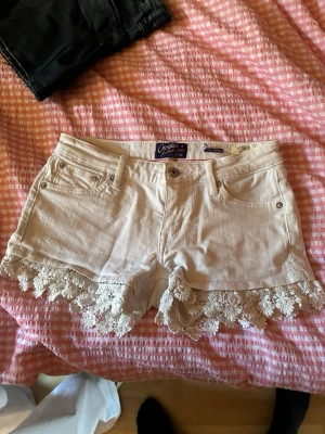 Vita shorts med spetsdetaljer - Säljer ett par vita shorts med snygga spetsdetaljer längs bensluten. Perfekta för sommarens alla äventyr och ger en romantisk touch till din outfit.