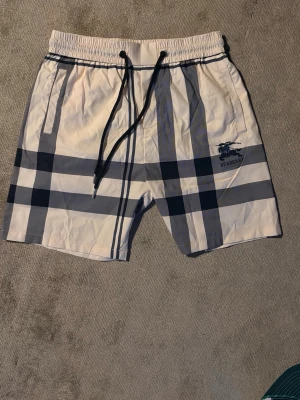Rutig shorts med snörning - Snygga shorts med klassiskt rutigt mönster i beige, svart och grått. Shortsen har elastisk midja med svart snörning och sidofickor. Tillverkade i ett lätt syntetmaterial som passar perfekt för sommaren.