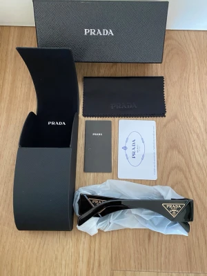 Svarta solglasögon från Prada - Snygga svarta solglasögon från Prada med rektangulär form och breda skalmar. Klassisk svart färg med guldfärgad Prada-logga på sidan. Kommer med originalfodral, putsduk och certifikat. Perfekt accessoar för en trendig look.