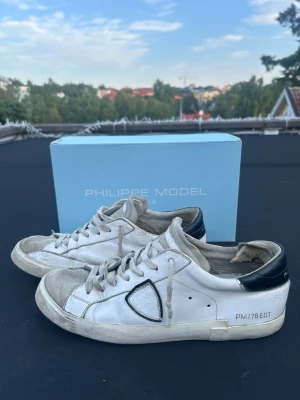 Vita Philippe Model sneakers - Säljer ett par vita Philippe Model sneakers med svarta detaljer på hälen och grå inslag på tån. Klassisk låg modell med vita snören och ikonisk sköldlogga på sidan. Snygg streetstil som passar till det mesta. 