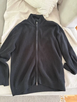 Svart ribbstickad ziptröja XL - Säljer en svart ribbstickad tröja med dragkedja framtill och hög krage. Tröjan har långa ärmar och är tillverkad i ett mjukt, tjockt material som passar perfekt när det är kyligt ute. Enkel och stilren design som funkar till det mesta.
