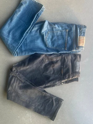 Två par jeans i blått och svart - Säljer två par jeans, ett par i klassiskt blå denim och ett par i svart tvättad denim. Båda har raka ben, fem fickor och normal midja. Perfekta för dig som vill ha en enkel och stilren look till vardags. Materialet är bomullsdenim. Köpt på deras hemsida nya.