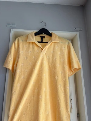 Gul kortärmad polotopp från H&M - Säljer en ljusgul polotopp från H&M i frotté med korta ärmar och krage. Toppen har ett diskret palm-mönster som ger en somrig vibe. Perfekt för dig som vill ha något bekvämt och stilrent till sommaren.