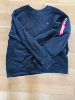 Svart sweatshirt från Alpha Industries - Cool svart sweatshirt från Alpha Industries med klassisk logga på bröstet och en röd tagg på ärmen. Tröjan har rund halsringning och ribbade muddar. 