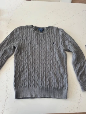 Grå kabelstickad tröja Polo Ralph Lauren - Kabelstickad grå tröja från Polo Ralph Lauren med rund halsringning och diskret broderad logga på bröstet. Tröjan är i XL-barnstorlek ca S i vanlig storlek. Använd endast fåtal gånger 
