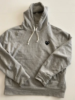 Cdg hoodie - Säljer en stilren grå hoodie med huva och vita dragsnören. På bröstet finns en liten svart hjärtdetalj med ögon. Hoodien har en klassisk passform och en stor ficka framtill. Perfekt för en chill och avslappnad look. 