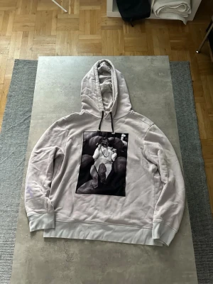 Limitato hoodie - Säljer en ljusbeige hoodie med stor svartvit fotoprint på bröstet. Hoodien har huva med svarta snören och breda muddar vid ärmar och nederkant. Materialet är mjukt och bekvämt, perfekt för en avslappnad stil.