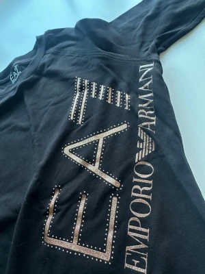 Svart sweatshirt EA7 Armani Äkta - Snygg svart sweatshirt från Emporio Armani med stora glittriga EA7-loggor och dekorativa nitar framtill. Tröjan har rund hals, långa ärmar och ribbade muddar vid ärmslut och nederkant. 