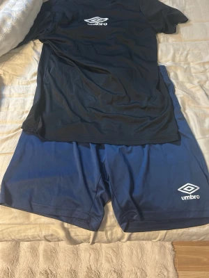 Svart Umbro t-shirt & blå shorts - Säljer ett sportset från Umbro med en svart t-shirt och ett par marinblå shorts. Båda plaggen har Umbro-loggan tryckt framtill och är tillverkade i ett lätt och snabbtorkande syntetmaterial, perfekt för träning eller fotboll.