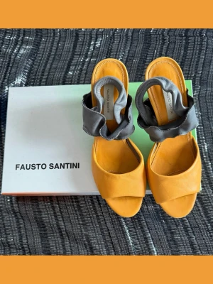 Gula sandaler från Fausto Santini - Unika sandaler i gult skinn från Fausto Santini med breda, vadderade grå remmar över vristen. Snygg öppen tå och stilren design som sticker ut. Perfekta för dig som vill ha något färgglatt och trendigt till sommaren.
