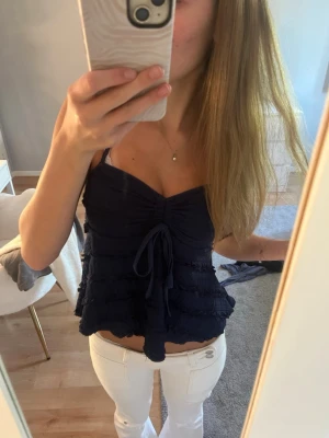 Mörkblå volangtop med knyt - Säljer en mörkblå topp med volanger och knyt framtill. Toppen är ärmlös med smala axelband och har en söt v-ringning. Materialet känns lätt och luftigt, perfekt för varma dagar. Knappast använd🥰