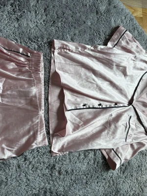 Rosa satinpyjamas från Censored XS - Snyggt tvådelat pyjamasset i glansigt rosa satin från Censored. Setet består av en kortärmad skjorta med svarta knappar och svarta detaljer samt matchande shorts med resår i midjan. Perfekt för mysiga kvällar hemma.