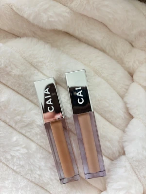Två CAIA concealers i neutrala toner - Säljer två CAIA concealers i snygga fyrkantiga förpackningar med silvriga lock. Ena concealern har en varm beige ton och den andra en ljusare neutral nyans. Perfekta för att täcka mörka ringar eller ojämnheter. Modern design och smidig applikator. Aldrig använda! 