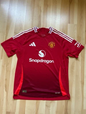 Manchester United matchtröja röd - Säljer en röd Manchester United matchtröja från Adidas med vita detaljer och klubbmärke på bröstet. Tröjan har Snapdragons logga på framsidan, vita ränder på axlarna och HEAT.RDY-material för bästa komfort. Perfekt för fotboll eller träning.