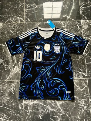 Argentina Adidas fotbollströja svart/blå - Snygg svart fotbollströja från Adidas med blått och vitt mönster, nummer 10 och Argentina AFA-emblem på bröstet. Tröjan har vita ränder på axlarna och FIFA World Champions 2022-märke. Perfekt för dig som älskar fotboll och vill sticka ut på planen.