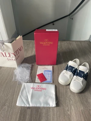 Valentino vita sneakers med blå detalj - Snygga vita sneakers från Valentino Garavani med bred blå läderdetalj över snörningen och guldfärgad logga på plösen. Klassisk låg modell i läder med vita skosnören och diskret design. Perfekta för dig som vill ha en exklusiv touch på din outfit.