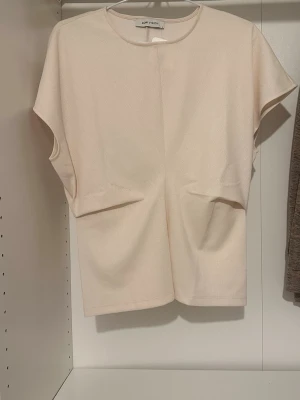 Ljusbeige kortärmad topp  - Minimalistisk ljusbeige topp  med rund hals och kortärmad design. Tillverkad i ett mjukt tyg som ger en clean och stilren look. Perfekt att matcha med jeans eller kjol för en enkel och snygg outfit.