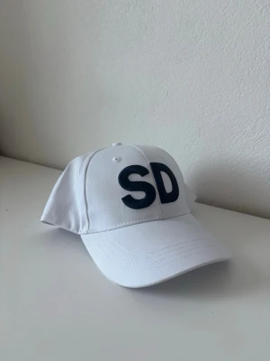 Vit keps med svarta bokstäver - Snygg vit keps med böjd skärm och justerbar rem bak. Framtill står stora svarta bokstäverna 'SD' broderat. Tillverkad i bomullstyg som känns lätt och bekvämt. Perfekt accessoar för en clean och stilren look.