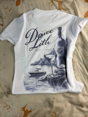 Vit t-shirt från Leon Marcel - Vit t-shirt från Leon Marcel med tryck på ryggen som visar en vinflaska, glas och båt i blå toner. Framsidan har en liten logga och texten 'Marcel' på bröstet. Klassisk passform och rund hals, perfekt för en avslappnad stil.