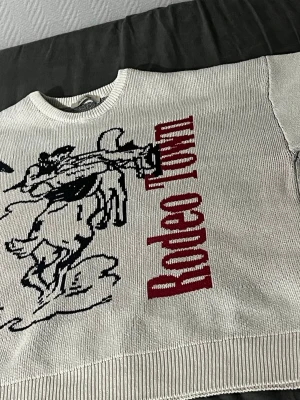 Vit tjock stickad tröja med tryck - Cool vit stickad tröja med svart cowboy-motiv och röd texten 'Rodeo Town' på framsidan. Tröjan har rund halsringning och ribbade muddar vid ärmslut och nederkant. Perfekt för dig som gillar streetwear och unika prints.