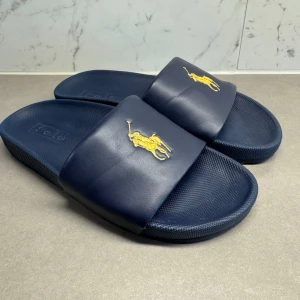 Marinblå slides från Polo Ralph Lauren - Snygga marinblå slides från Polo Ralph Lauren med klassisk gul logga på remmen. Tillverkade i syntetmaterial med bekväm sula och halksäker undersida. Perfekta för sommaren och enkel att matcha till olika outfits.