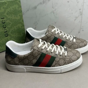 Gucci Ace sneakers med GG-mönster - Snygga Gucci Ace sneakers i beige canvas med klassiskt GG-mönster och gröna/röda sidoremsor. Skorna har vita snören, vit sula och detaljer i mörkgrönt vid hälen. Perfekta för dig som vill ha en ikonisk och trendig look.