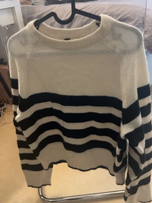 Randig stickad tröja från H&M Divided - Stickad tröja från H&M Divided i vitt och svart med breda horisontella ränder. Tröjan har rund halsringning, långa ärmar och ribbade muddar. Perfekt för en avslappnad och trendig look.