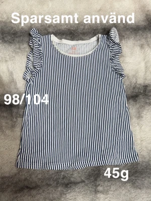 Randig blåvit linnetopp med volang - Söt ärmlös topp med smala blå och vita ränder och volangdetaljer på axlarna. Rund halsringning och lätt, luftig känsla. Perfekt för varma dagar och enkel att matcha till olika outfits.🌸Sparsamt använd. Storlek 98/104