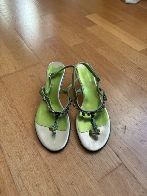 Gröna sandaletter med pärldetaljer - Säljer ett par glansiga sandaletter i metallicgrönt med tunna remmar och små pärlor längs t-remmen. Sandalerna har öppen tå och låg klack, samt justerbart spänne vid ankeln. Perfekta för dig som vill sticka ut med färg och detaljer. Aldrig använda endast testade. 