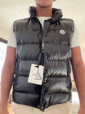 Svart dunväst från Moncler - Snygg svart dunväst från Moncler med hög krage och dragkedja framtill. Västen har två sidofickor med dragkedja och Moncler-logga på bröstet. Tillverkad i glansigt material som ger en modern look. Perfekt för lager-på-lager-stil.