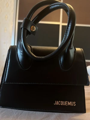Svart Jacquemus Le Chiquito väska - Säljer en svart Jacquemus Le Chiquito handväska i slätt läder med guldfärgad logga framtill. Väskan har ett topphandtag med dekorativ knäppning och en stilren, strukturerad form. Perfekt accessoar för att lyfta din outfit. Kommer med en kedja för om man vill bära den som en crossbody