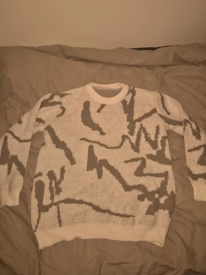 Vit stickad tröja med grått mönster - Säljer en vit stickad tröja med abstrakt grått mönster. Tröjan har rund halsringning, långa ärmar och ribbade muddar vid ärmslut och nederkant. Perfekt för dig som gillar unik design och vill ha något som sticker ut i garderoben.