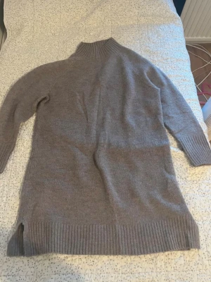 Ljusgrå stickad polotröja - Mysig ljusgrå stickad polotröja med lång passform och ribbade muddar vid ärmslut, nederkant och krage. Tröjan har en hög, bred polokrage och små slitsar i sidorna. Perfekt för kyliga dagar och enkel att styla till olika outfits.