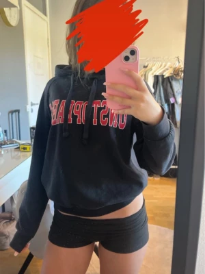 Svart hoodie - Säljer en svart cropped hoodie med huva och dragsnören. Framtill finns ett stort tryck i rött och vitt. Hoodien har en avslappnad passform och ribbade muddar vid ärmarna. Perfekt för en chill och sportig look. Storlek XXS från Gina Tricot men passar även xs. Lite knottrig men inget man tänker på.