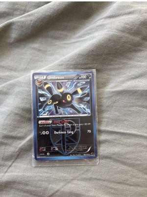 Pokémonkort Umbreon Darkness Fang - Säljer ett Pokémonkort med Umbreon, motivet visar Umbreon med blå och gula detaljer. Kortet har attacken Darkness Fang och HP 100. Det är inplastat för extra skydd och har en snygg, glansig yta. Perfekt för samlare eller Pokémonfans!