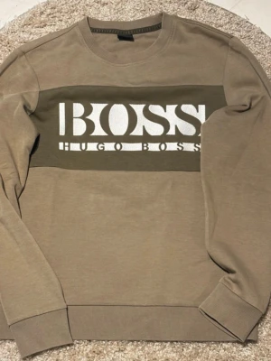 Beige sweatshirt från Hugo Boss - Snygg beige sweatshirt från Hugo Boss med stort vitt logotryck över bröstet och kontrasterande mörkare panel. Den är storlek xs men skulle säga den passar s också den är helt ny inga skador eller solbränna på den eller något sånt knappt använd pris kan diskuteras och ge byten beror på vad det är och den säljs inte längre 