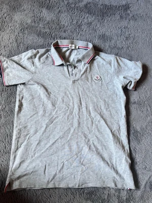 Grå pikétröja från Moncler - Snygg grå pikétröja från Moncler med klassisk krage och knappar framtill. Tröjan har Moncler-logga på bröstet och detaljer i rött, vitt och blått på krage och ärmslut. Perfekt till en clean och sportig stil. STORLEK LARGE MEN PASSAR SOM SMALL
