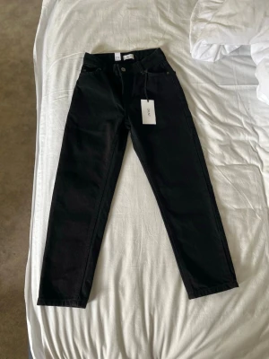 Svarta raka jeans med hög midja - Säljer ett par svarta jeans med rak passform och hög midja. Jeansen har klassiska fem fickor och bälteshällor. Materialet är kraftigt denim som ger en snygg siluett. Perfekta till en avslappnad eller trendig look.