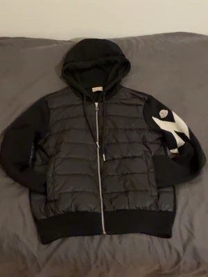 Svart pufferjacka med huva från Moncler - Snygg svart pufferjacka från Moncler med huva och dragkedja framtill. Jackan har vadderad kropp, stickade ärmar och ribbade muddar. På ena ärmen finns Moncler-logga och ett vitt mönster. Perfekt för kalla vinterdagar.