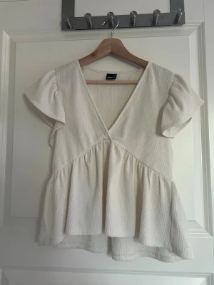 Vit v-ringad topp från Gina Tricot - Säljer en ljus topp från Gina Tricot i storlek S. Toppen har v-ringning, korta ärmar och volang nedtill för en luftig och söt look. Perfekt till jeans eller kjol för en fräsch stil.