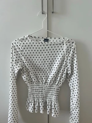 Prickig topp från Gina Tricot - Supersöt vit topp från Gina Tricot med svarta prickar, långärmad och smockad midja som ger en snygg siluett. Toppen har en lätt volang nedtill och är gjord i ett stretchigt, ribbat material. Perfekt för dig som gillar trendiga detaljer och mönster!