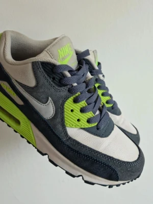 Nike Air Max 90 sneakers grå och lime - Nike Air Max 90 sneakers i grått, vitt och limegrönt. Klassisk chunky sula med synlig Air-enhet, snörning och ikonisk Swoosh på sidan. Ovandel i en mix av mocka och syntetmaterial. Perfekta för dig som vill sticka ut med färg och stil.