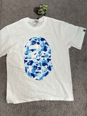 Vit BAPE t-shirt med blått camomönster - Säljer en clean vit t-shirt från BAPE med det ikoniska apansiktet i blått camomönster både fram och bak. Klassisk passform och rund hals. Perfekt för dig som gillar streetwear och vill sticka ut lite extra. TAGGEN EJ KVAR