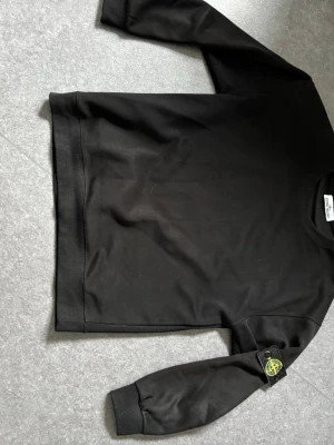 Svart sweatshirt från Stone Island - Svart sweatshirt från Stone Island med klassisk logotyp-patch på vänster ärm. Tröjan har rund hals, ribbade muddar och är tillverkad i mjukt bomullsmaterial. Perfekt för en stilren och avslappnad look.
