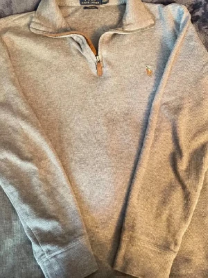 Ralph lauren quarter zip - Snygg grå stickad tröja från Ralph Lauren med halv dragkedja och klassisk krage. Tröjan har lång ärm och den ikoniska broderade loggan på bröstet. Perfekt till jeans eller chinos för en clean och stilren look.