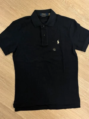 Marinblå pikétröja från Polo Ralph Lauren - Klassisk svart pikétröja från Polo Ralph Lauren med korta ärmar och krage. Tröjan har knappar framtill och den ikoniska broderade loggan i vitt på bröstet. Perfekt till jeans eller shorts för en clean och stilren look.