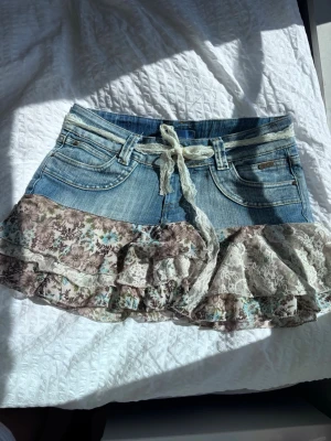 Kort jeanskjol med volanger och spets - Söt kort jeanskjol med volanger i blommigt tyg och spetsdetaljer. Kjolen har en ljusblå denimtopp med fickor och ett knytband i spets i midjan. Volangerna är i beige, brunt och blått med blommigt mönster och spetsinslag för en romantisk vibe. Fet är storleken medium men den funkar väldigt bra för small också!